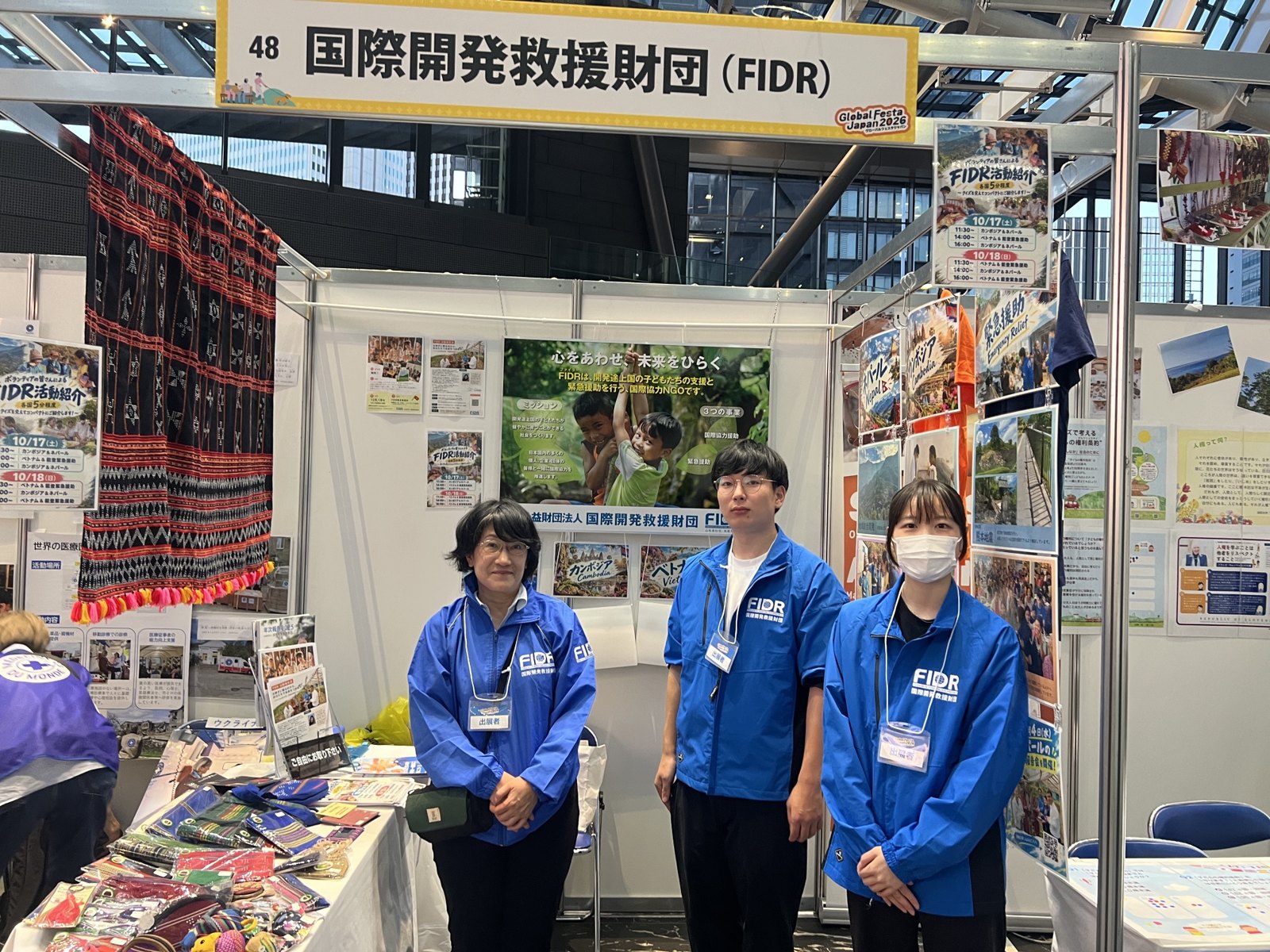 グローバルフェスタJAPAN 2026のFIDRのブース。青いジャケットのスタッフ3人が活動パネルの前に並ぶ