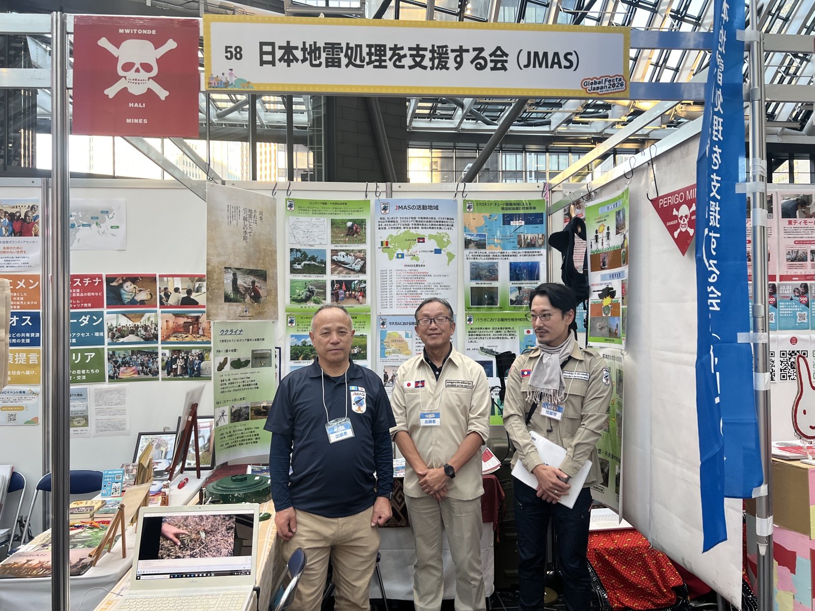 グローバルフェスタJAPAN 2026のJMASのブース。活動地域のパネルの前にスタッフ3人が並ぶ