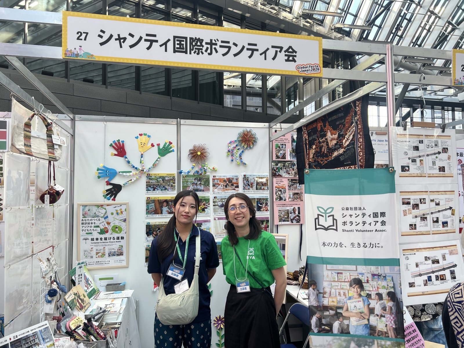 グローバルフェスタJAPAN 2026のシャンティ国際ボランティア会のブース。スタッフ2人が絵本を届ける運動のパネルの前に立つ