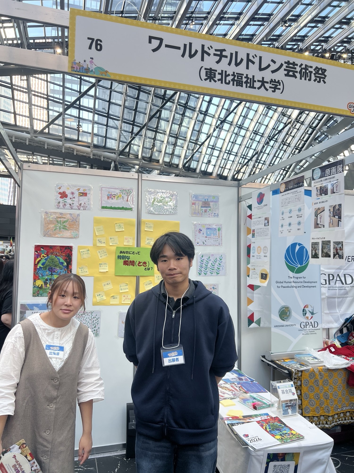 グローバルフェスタJAPAN 2026のワールドチルドレン芸術祭のブース。世界の子どもたちの絵の前に学生2人が立つ